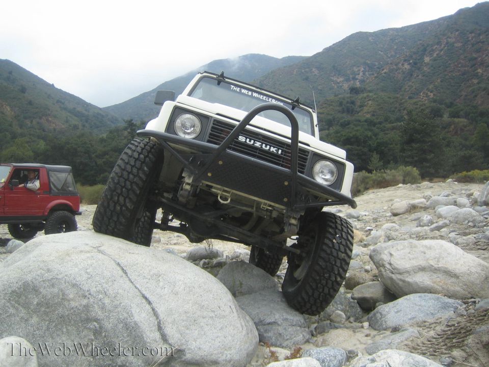 White 1987 Suzuki Samurai climbing over rocks on a trail, red Suzuki Samurai in background