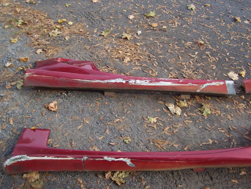 Two red car side skirts lying on the ground, showing damage and wear