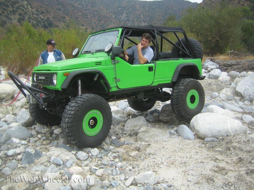 Green 1987 Suzuki Samurai with large tires and a roll cage, parked on rocks