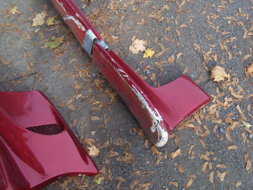 Damaged red car fender and side skirt on asphalt with fallen leaves. White primer shows through chipped paint