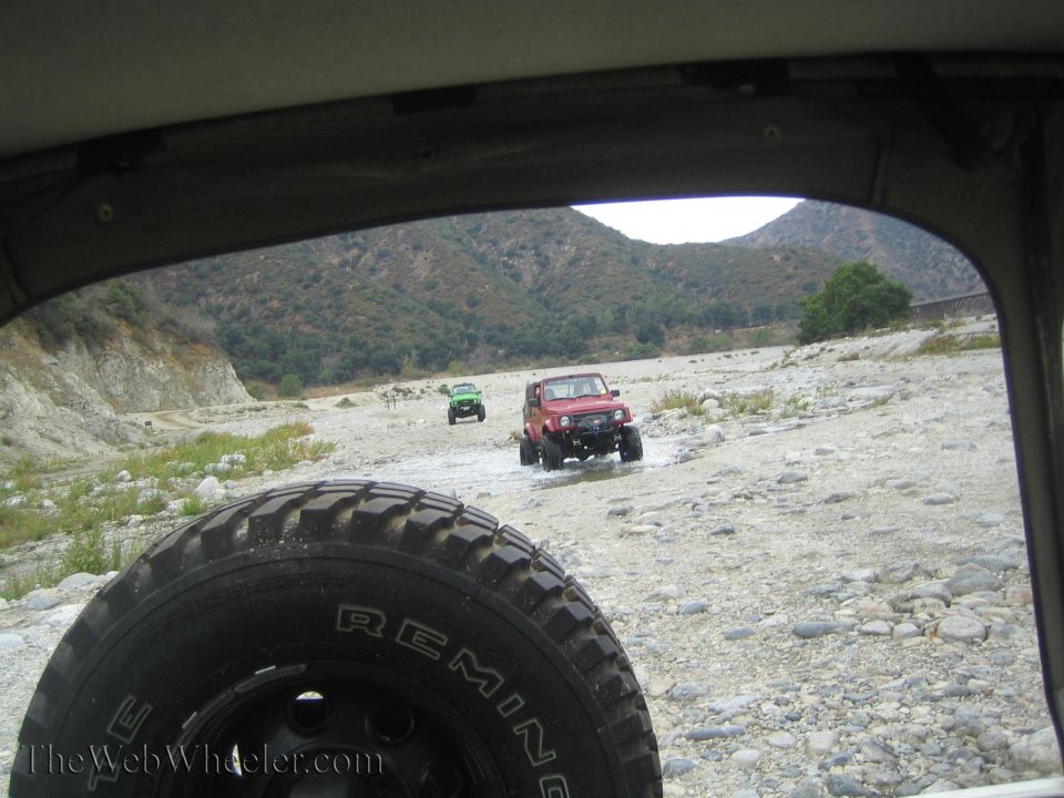Red 1987 Suzuki Samurai driving through a rocky riverbed with a green Jeep behind it