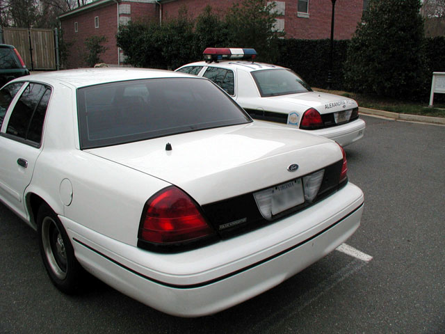 White Crown Victoria from rear three-quarter with an Alexandria Police cruiser parked beyond