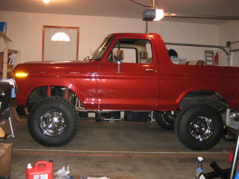 1979 Ford Bronco - Side profile of a red Ford Bronco, modified with a lift kit and custom wheels, inside a garage.