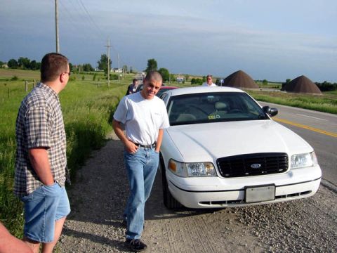 1998 Ford Crown Victoria - A white 1998 Ford Crown Victoria is parked on the side of a rural road with two men standing nearby.