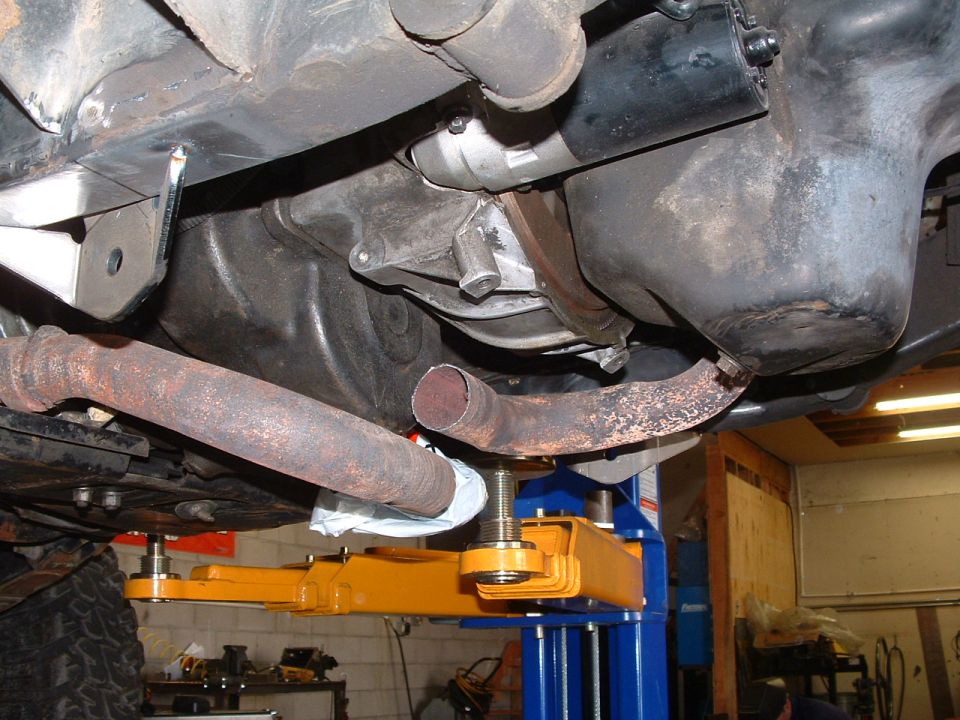 Underside of a vehicle on a lift, showing exhaust pipes, starter motor, and oil pan
