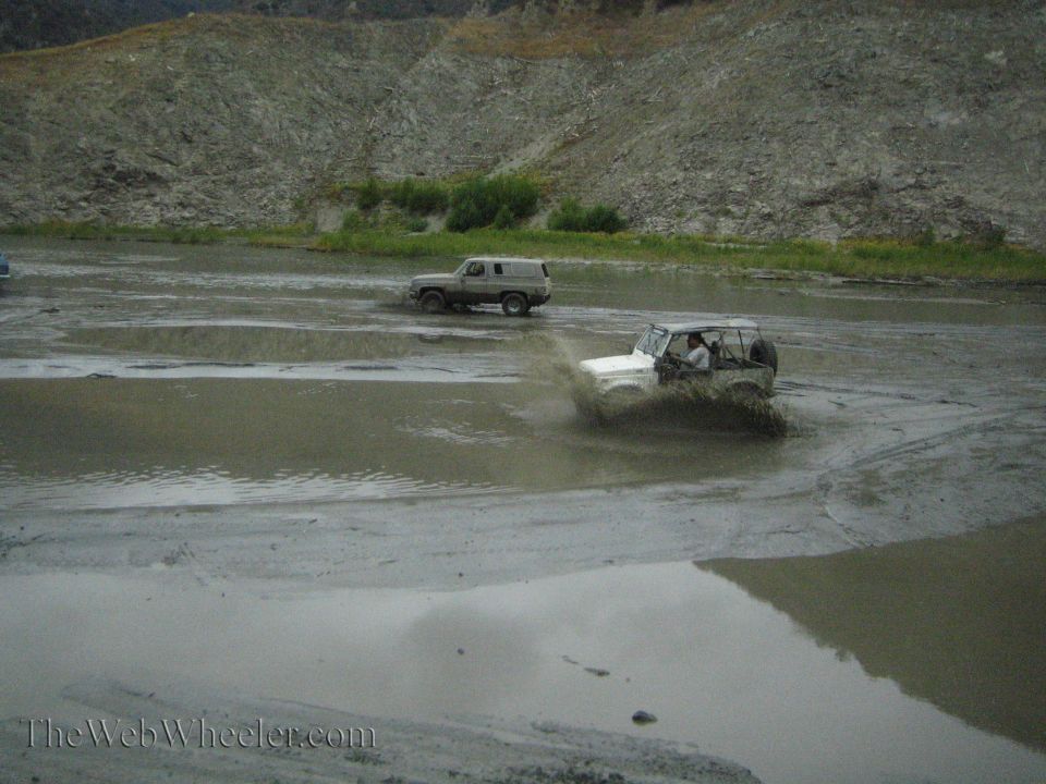 White off-road vehicle splashing mud, beige SUV in background, muddy water, rocky hill