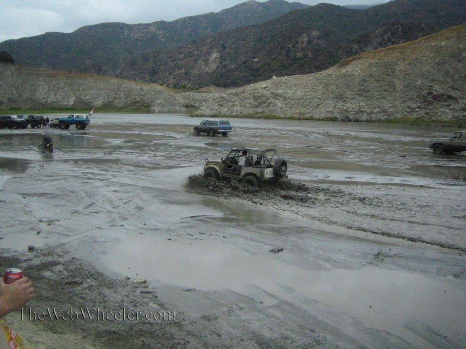 Jeep-like vehicle splashing through mud with other trucks and SUVs in the background near a lake and hills