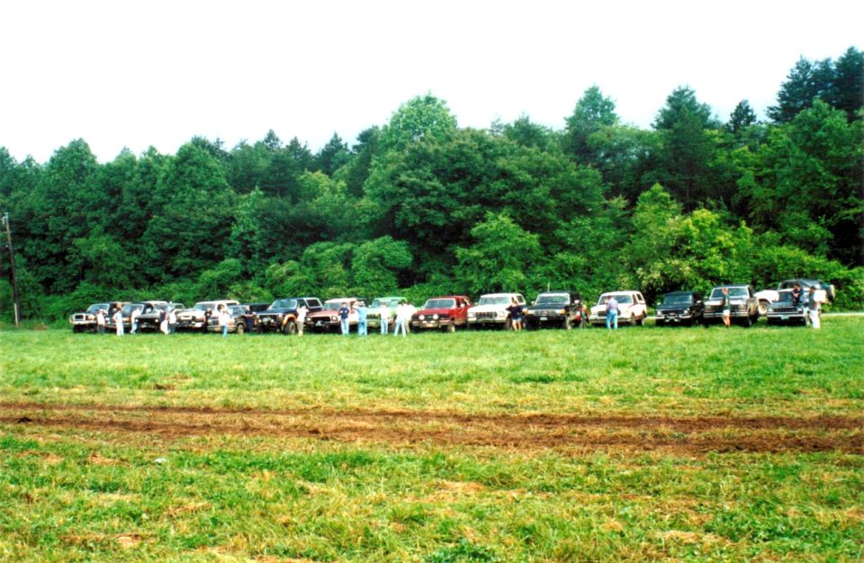 Row of trucks and SUVs with people lined up along a grassy field by the woods