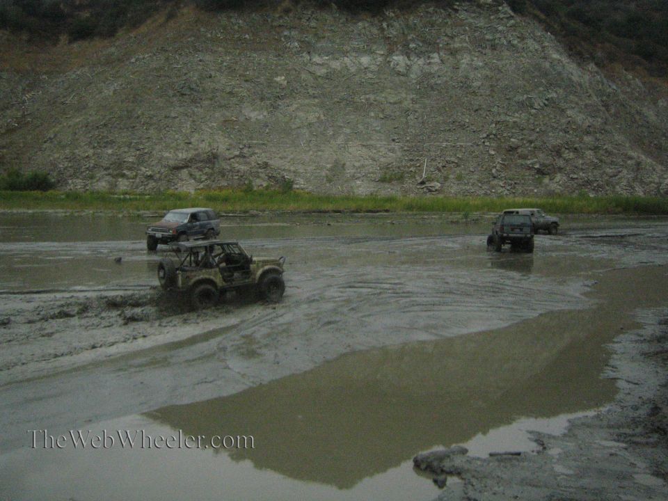 Several off-road vehicles driving through a muddy area with a rocky hill in the background