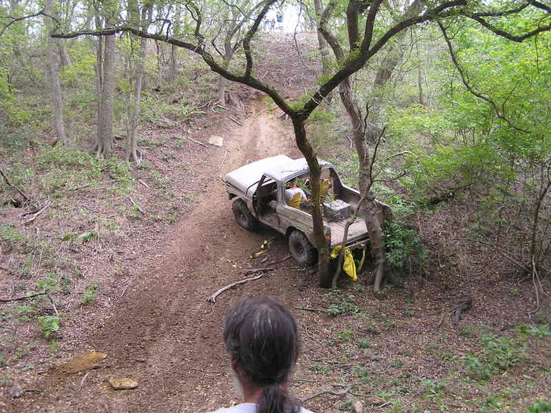 Tan 4x4 with crushed roof stuck against a tree at the base of a steep wooded dirt hill, recovery straps nearby
