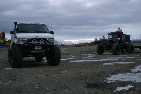 2003 Jeep TJ - Two vehicles, a white van and a black off-road machine, sit parked in a desolate landscape under a cloudy sky.