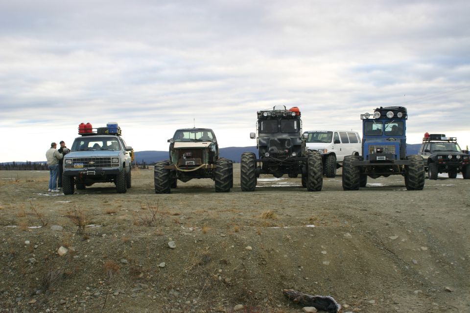 Four modified vehicles parked on a dirt lot under a cloudy sky. Two people stand near the front left vehicle
