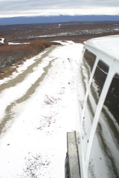 2003 Jeep TJ - A white van travels down a snowy, unpaved road with barren trees and distant mountains under a cloudy sky.
