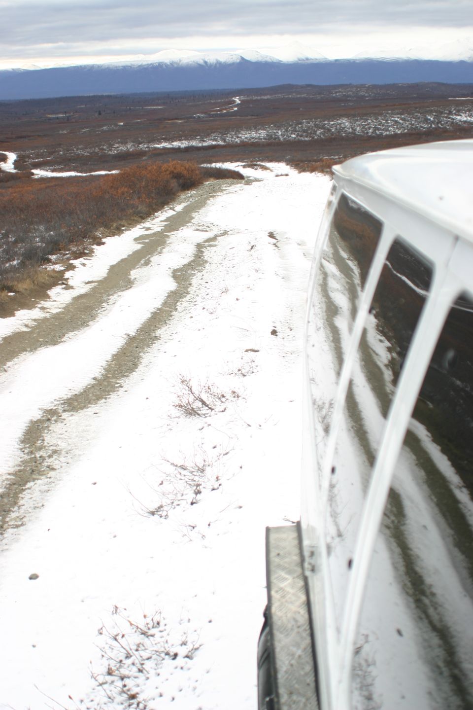 Side view of white van driving on a dirt road with snow and brush, mountains in distance