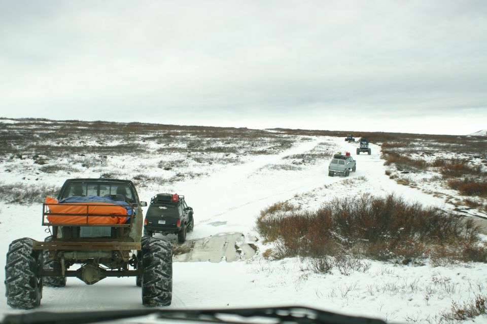 Truck with oversized tires and orange cargo in the bed, driving on a snowy path with other vehicles