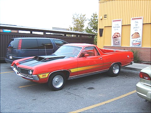 1967 Ford Ranchero - A red 1970 Ford Ranchero is parked next to a blue van and a silver car in front of a yellow building.