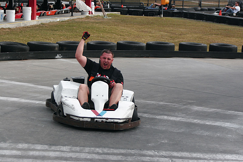 Man in a white go-kart with arms raised on a track, other go-karts visible in background