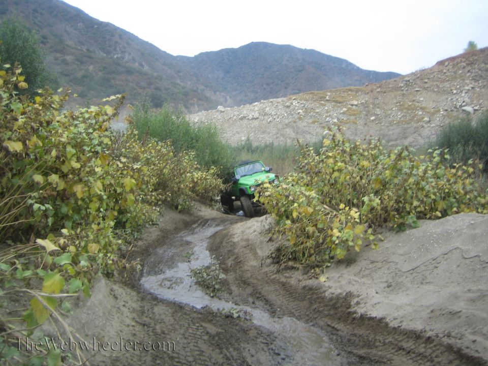 Green 1987 Suzuki Samurai driving on a muddy, overgrown trail