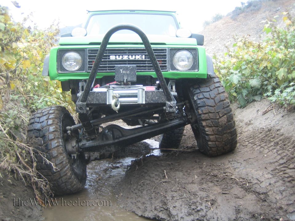 Green 1987 Suzuki Samurai with a winch and brush guard driving through mud