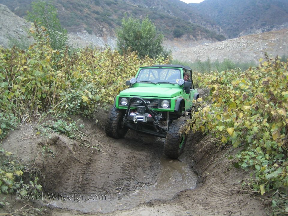 Green 1987 Suzuki Samurai off-roading through mud, front view