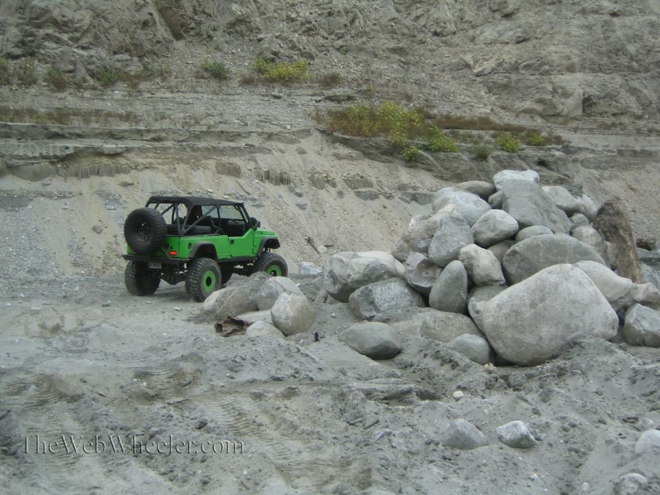 Green off-road vehicle with a spare tire on the back, parked on a dirt road next to a pile of rocks