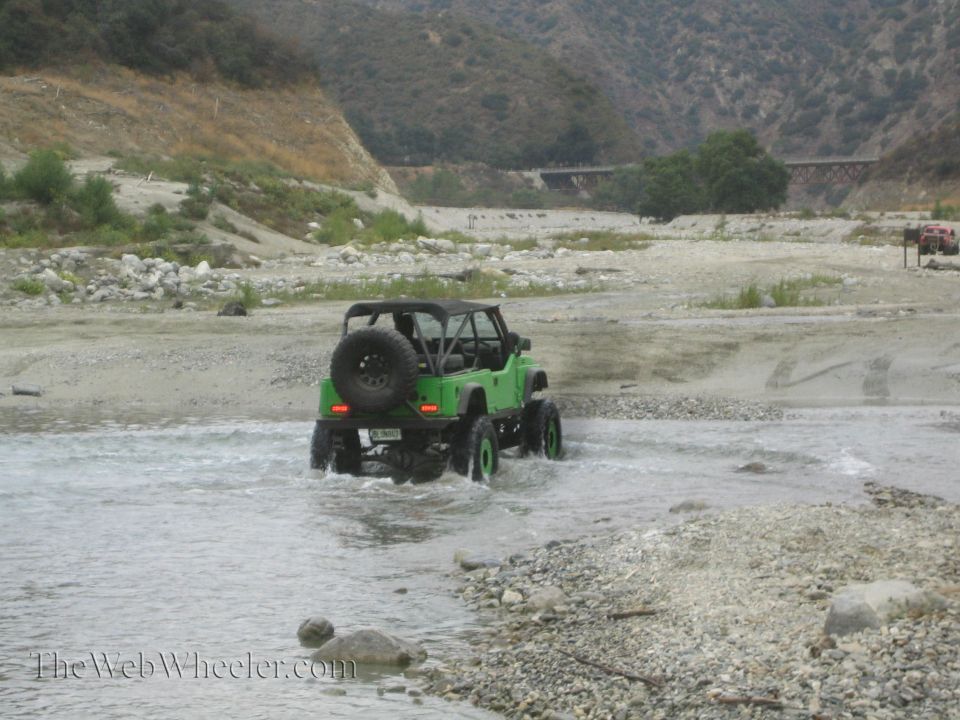 Green off-road vehicle with spare tire on back driving through shallow water in a rocky riverbed. Another red vehicle is visible in the distance