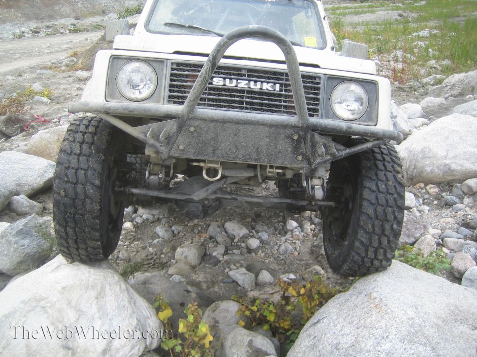 Front view of a white 1987 Suzuki Samurai with a custom bumper, navigating large rocks on a trail