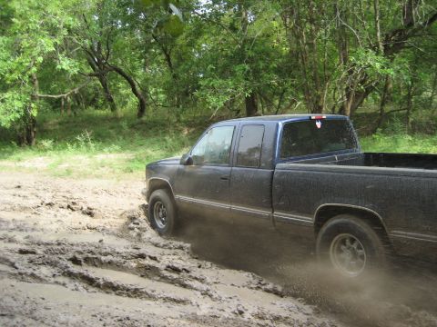 1986 Ford Bronco - A dark blue pickup truck navigates a challenging muddy path surrounded by trees.
