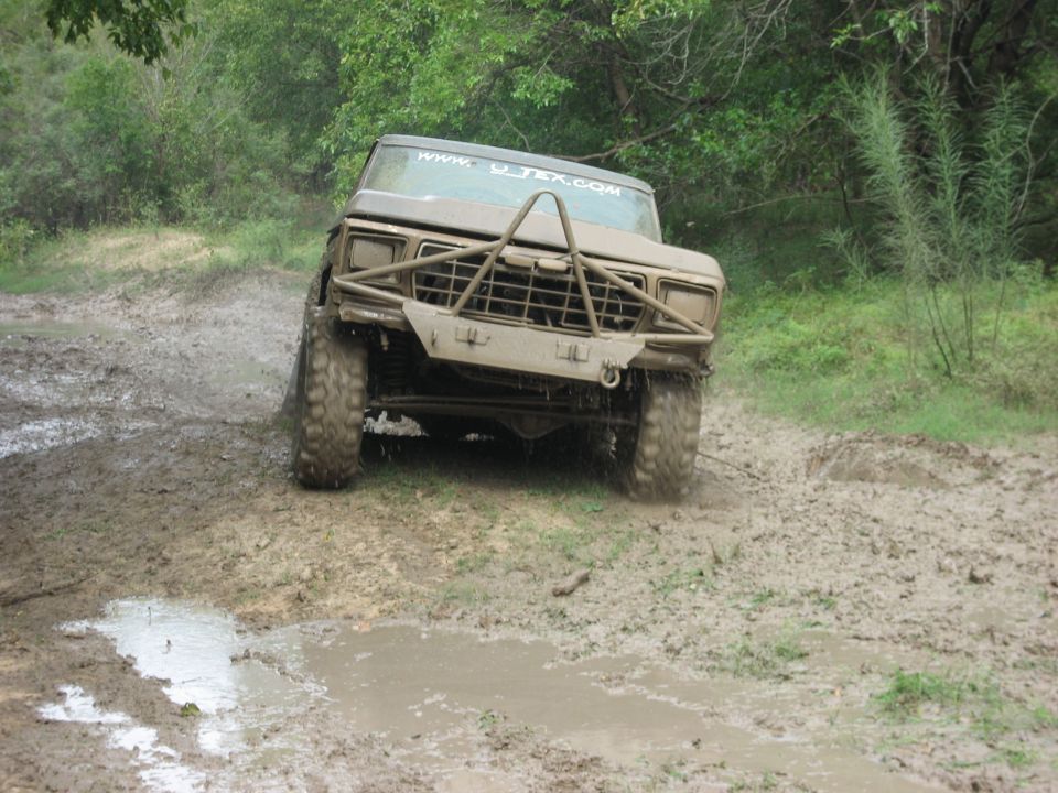 Mud-covered 1980s Ford Bronco with aftermarket bumper driving through mud