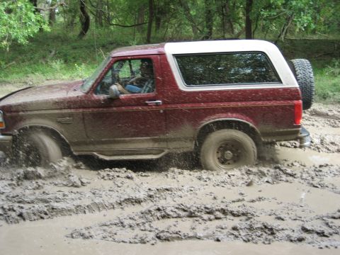 1986 Ford Bronco - A red SUV navigates through a muddy off-road trail, with one wheel deep in the mire.