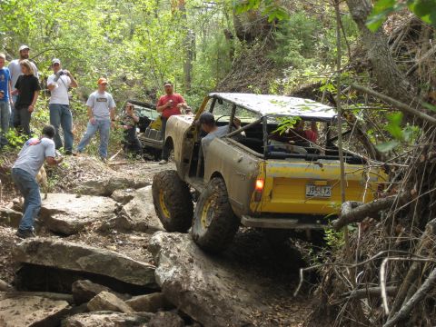 1986 Ford Bronco - A group of people watch as a modified off-road vehicle tackles a challenging rocky incline in a wooded area.
