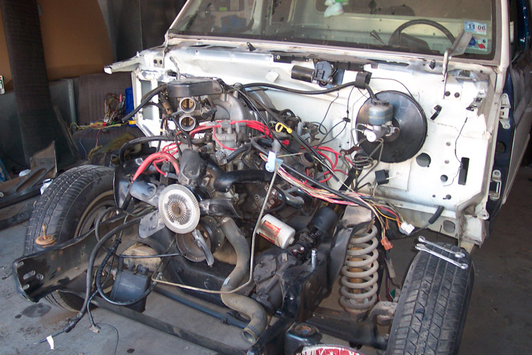 White pickup with front end removed showing exposed engine, wiring, and suspension during teardown in a garage