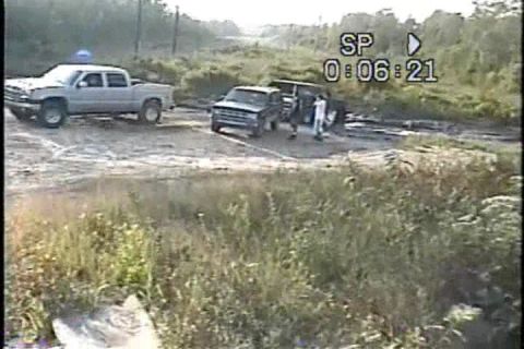 1989 Ford Bronco - Two trucks are in a dirt lot with two people standing between them, possibly engaged in some activity.