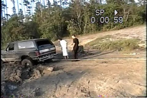 1989 Ford Bronco - A 1989 Ford Bronco is stuck in a muddy off-road situation with two individuals observing.