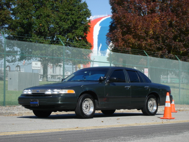 Dark green Ford Crown Victoria sedan with lace wheels parked by a chain-link fence