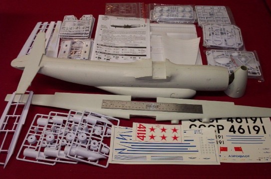 Model airplane kit parts and decals laid out on a red surface. Includes fuselage, wings, and sprues with small components
