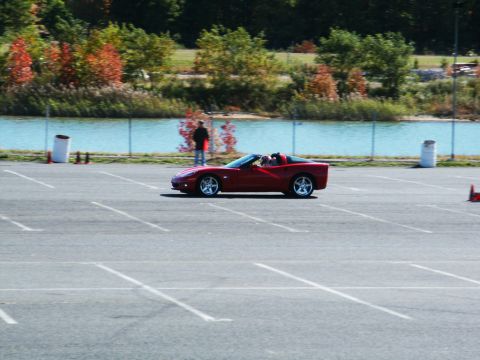2001 Mercury Grand Marquis - A red convertible sports car is seen on a paved lot near a body of water, with trees and a person in the distance.