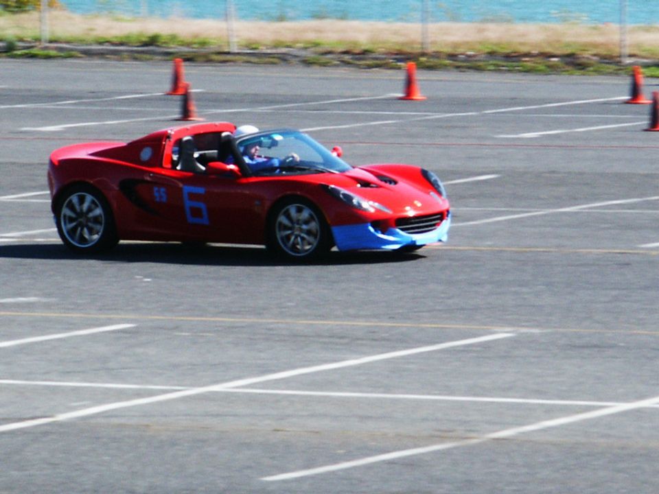 Red sports car with blue front bumper and side number 6, on a paved lot with orange cones