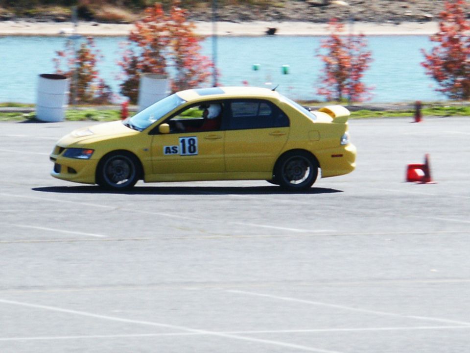 Yellow sedan with racing numbers 'AS 18' on the door, driving on a track with cones and a lake in the background