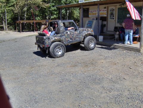 1978 Ford Bronco - A camouflage-wrapped off-road vehicle is parked on a gravel lot near a building with several people present.
