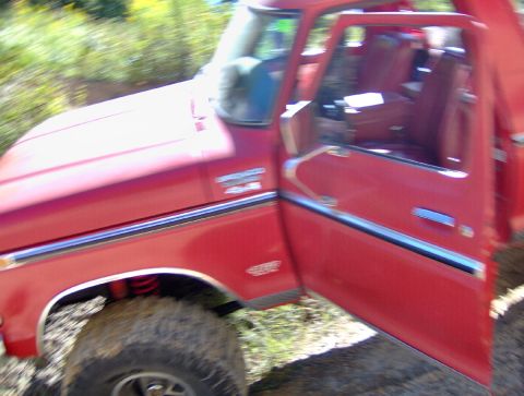 1978 Ford Bronco - A red 1978 Ford Bronco is seen from a 3/4 front angle with the driver's door open, revealing its interior and a muddy tire.