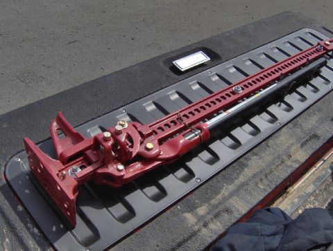 1978 Ford Bronco - A freshly painted red high-lift farm jack with gold-plated hardware lies on a black ribbed body panel or bumper cover in the sun. The jack'…