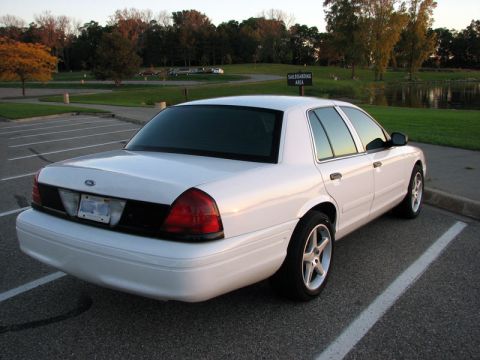 2003 Ford Crown Victoria - Three-quarter rear view of a white Ford Crown Victoria sedan parked in an empty lot at golden hour, showing dark-tinted rear glass, a black…