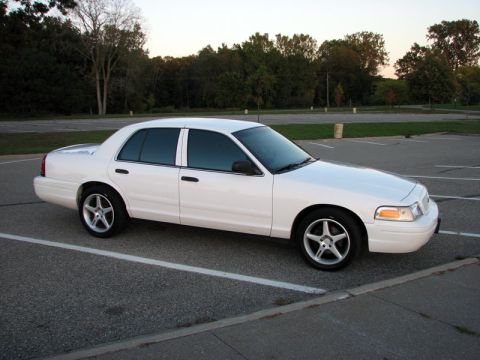 2003 Ford Crown Victoria - A white Ford Crown Victoria sedan photographed from the passenger side in an empty parking lot at golden hour, wearing dark-tinted windows,…