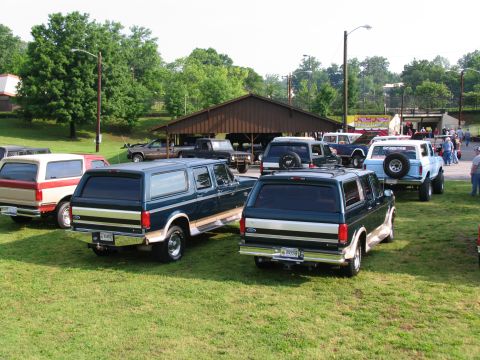 1995 Ford C-150 (Centurion Conversions) - A dark green 1995 Ford Bronco with a tan stripe and chrome trim is parked on grass alongside other vehicles.