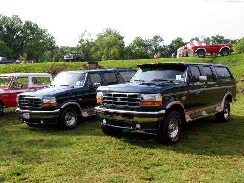 1995 Ford C-150 (Centurion Conversions) - Two Ford Broncos are parked on a grassy field with trees and a tent in the background.