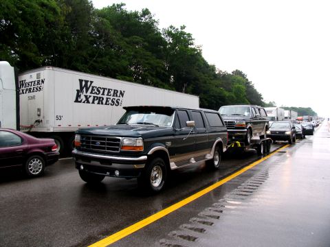 1995 Ford C-150 (Centurion Conversions) - Two Ford Bronco SUVs are seen on a highway, one towing the other on a trailer.