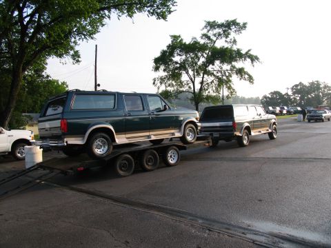 1995 Ford C-150 (Centurion Conversions) - A dark SUV is towed by a green and tan Ford Bronco, with other vehicles parked in the background.