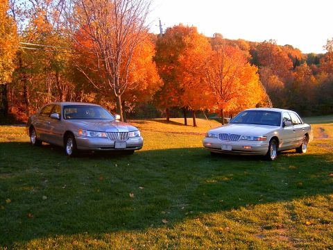 1996 Mercury Grand Marquis - Two silver full-size sedans parked on a grassy lawn in front of vivid orange and red autumn trees; a Lincoln Town Car on the left and a Mer…