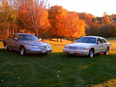 1996 Mercury Grand Marquis - Two full-size sedans parked on a grassy lawn with vivid autumn foliage in the background; a silver Lincoln Town Car on the left and a light…